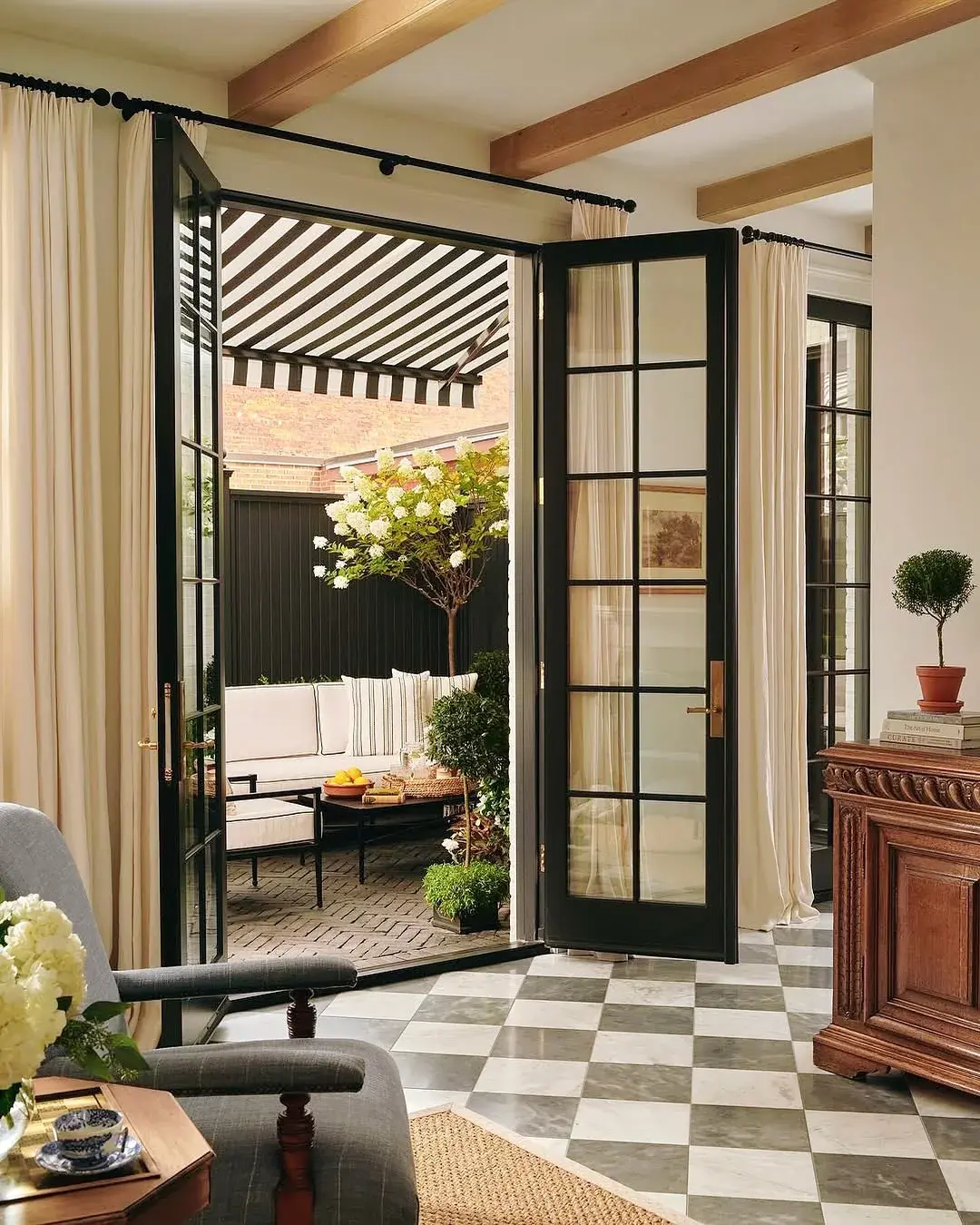 Cream curtains on a black rod hung wide of open black French doors above a grey and white marble checkerboard floor