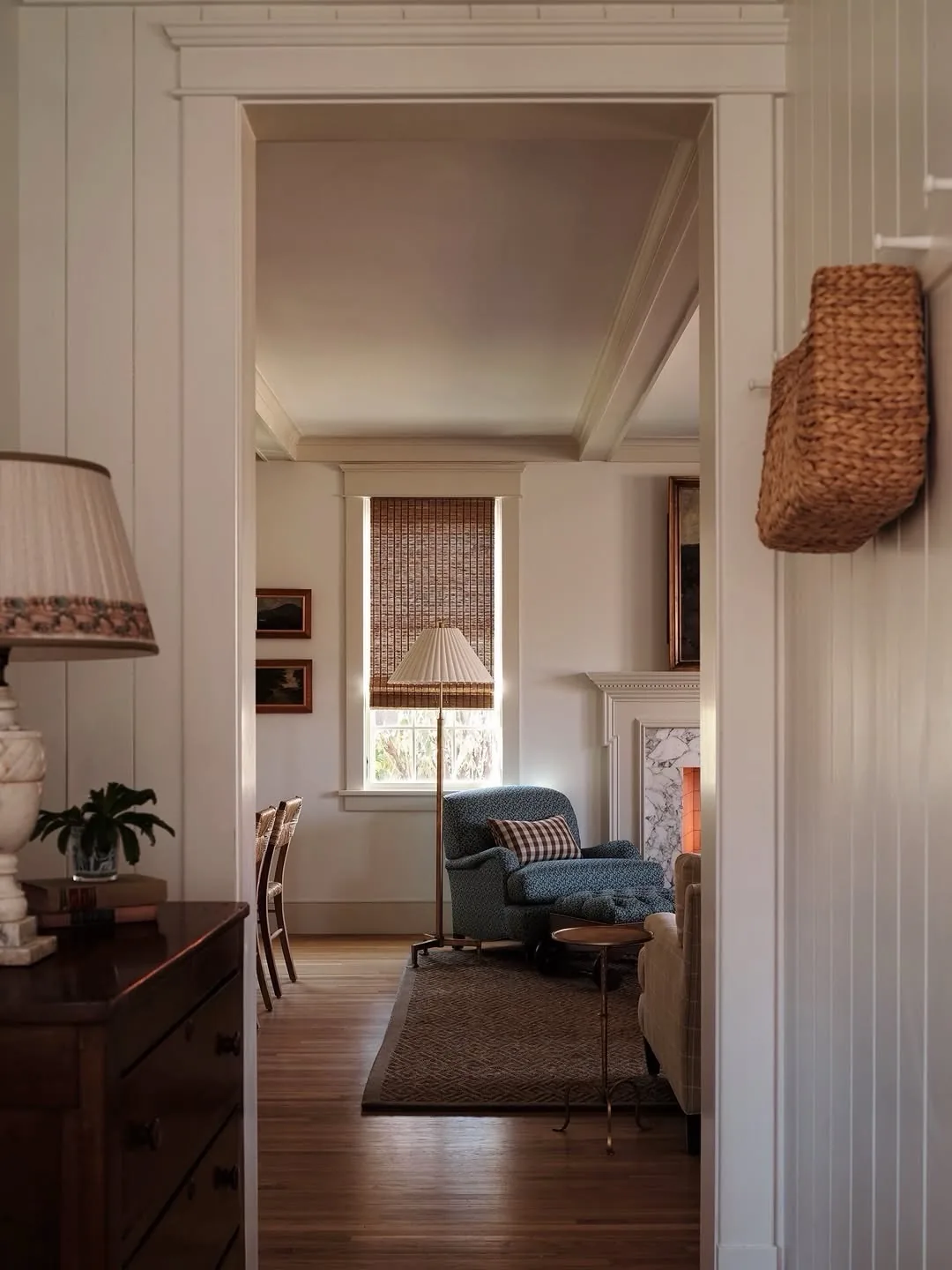 View through a panelled doorway to a blue armchair, a lit fire and a woven blind in a small sitting room