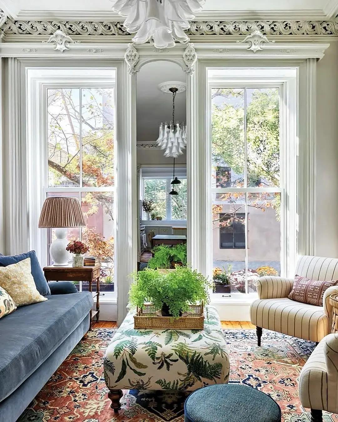 Tall room with a deep plaster cornice, a blue velvet sofa and a fern-print ottoman between two sash windows
