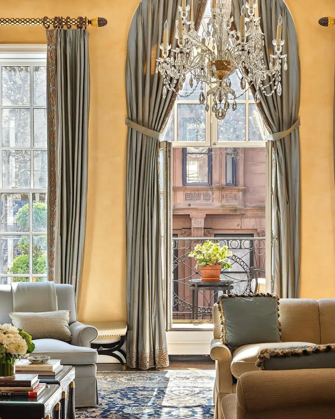 Grey blue curtains with an embroidered border held back on a carved rod beside a crystal chandelier