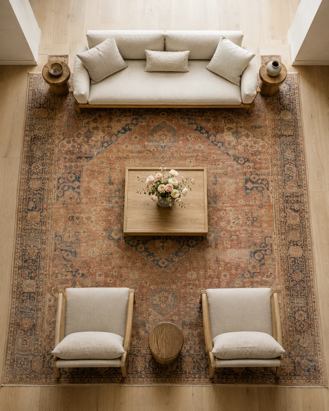 Overhead view of a faded antique Persian rug anchoring a cream linen sofa and two sand lounge chairs on wide oak floors